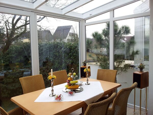 Heller Wintergarten mit Esstisch, Korbstühlen und Ausblick auf Garten und Häuser.